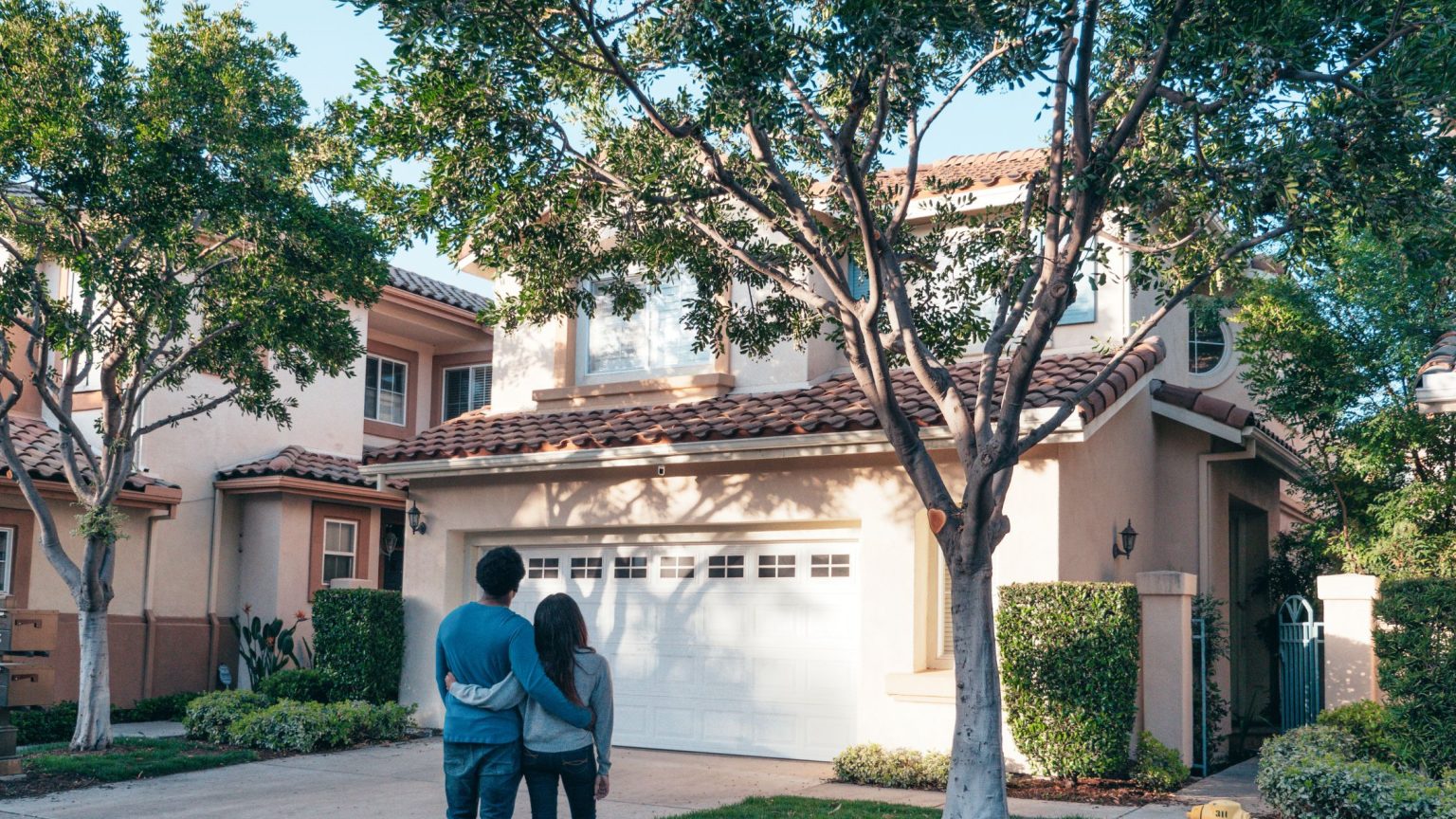 Couple standing arm in arm in front of a two-story stucco home with a tree-lined driveway